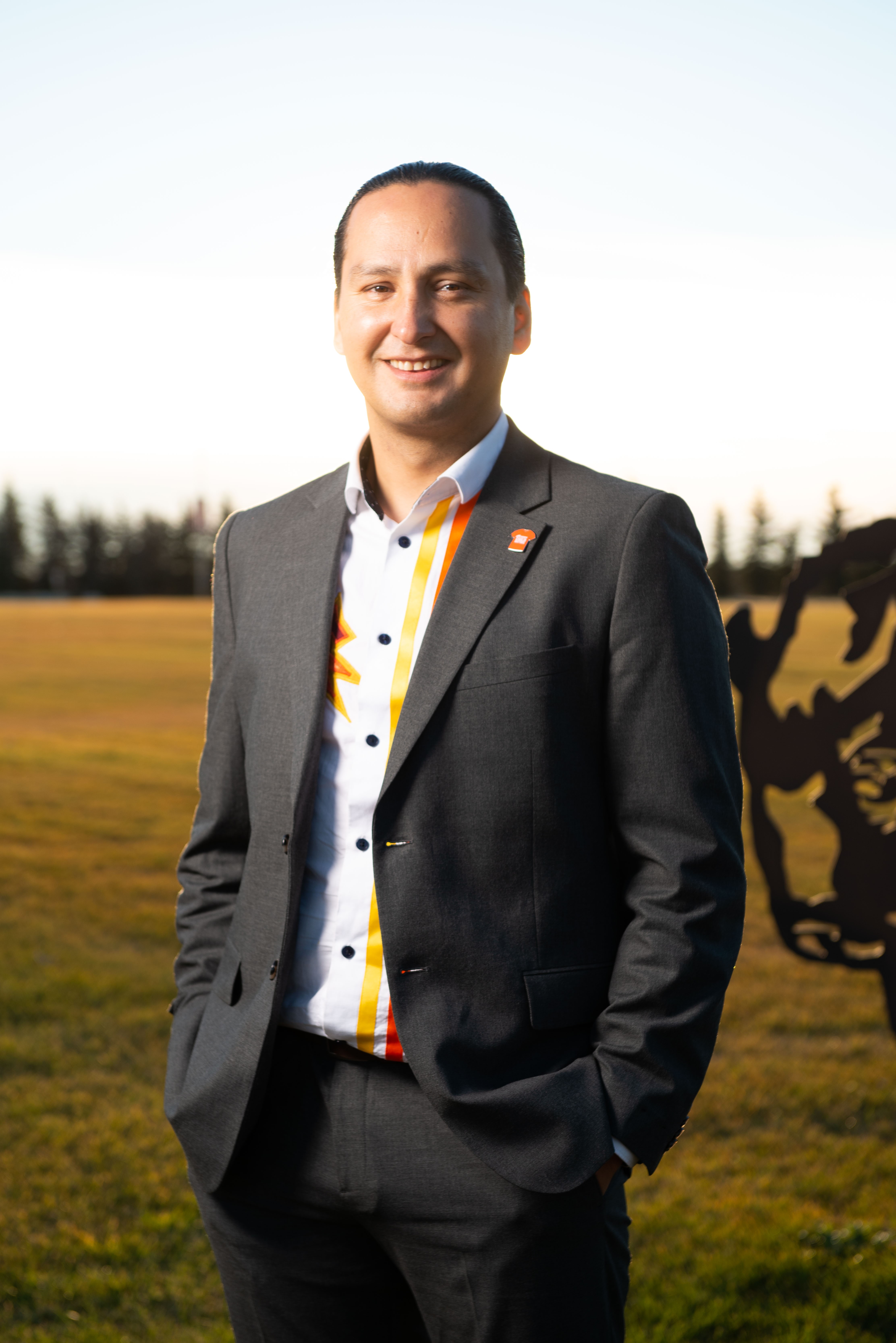Cadmus Delorme stands outside wearing a black suit and a white patterned shirt. His hands are in his pockets and he smiles at the camera. His dark hair is pulled back away from his face.