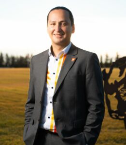 Cadmus Delorme stands outside wearing a black suit and a white patterned shirt. His hands are in his pockets and he smiles at the camera. His dark hair is pulled back away from his face.