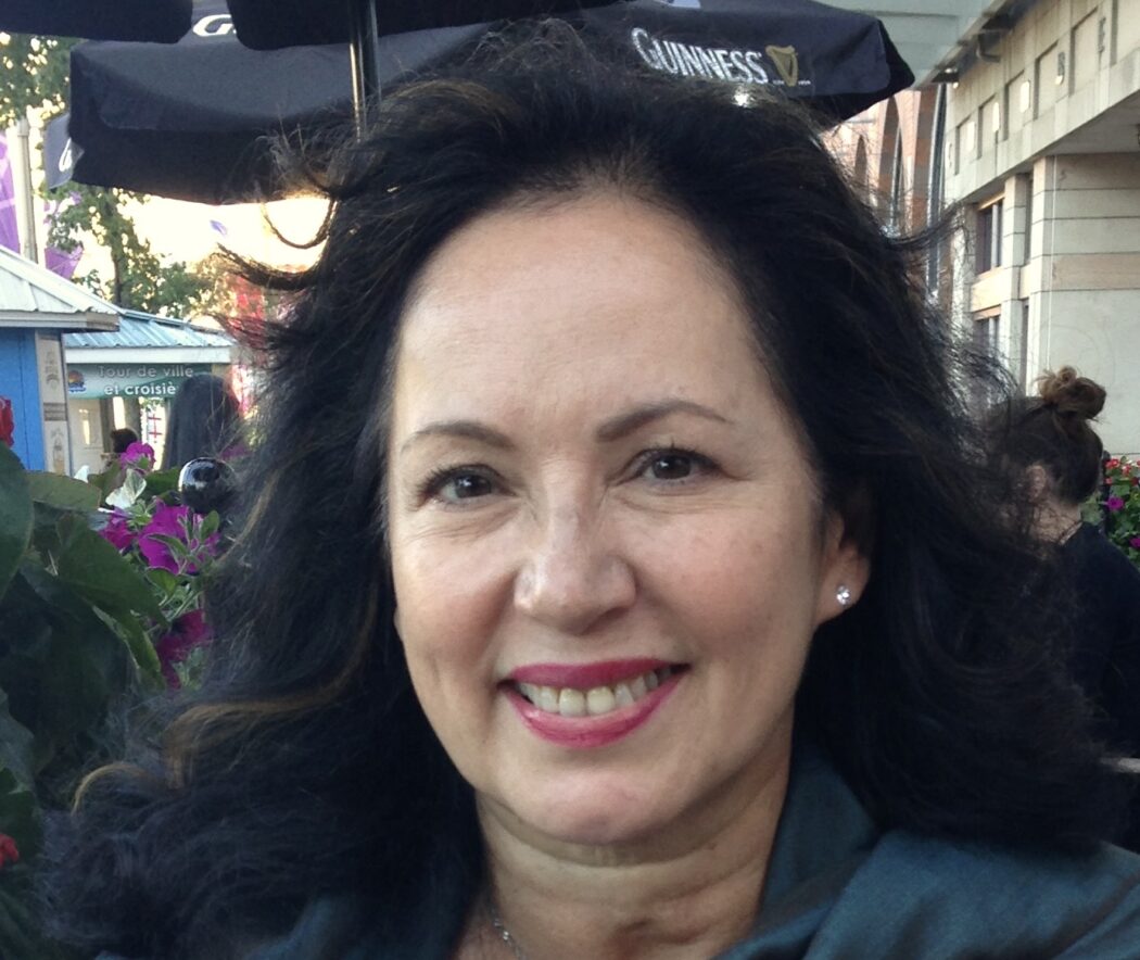 Deborah Pelletier has black hair that falls around her shoulders. She is wearing a white shirt with a silky turquoise scarf draped over her shoulders.