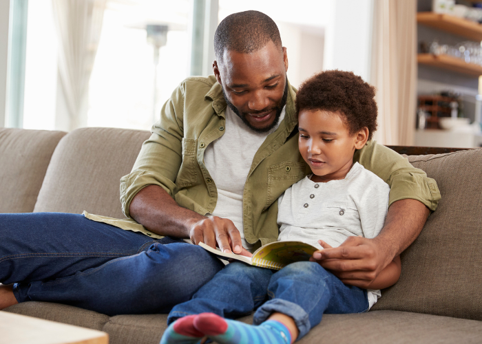 A father sits with his son on a couch. They are reading a book together.