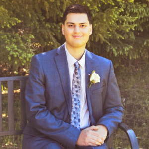 Jaydan Harrison wears a dark blue suit with a white shirt and a patterned tie. He has black hair and dark eyes and smiles at the camera.