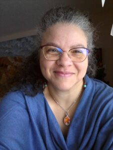 Jennifer S. Wallace has curly black and grey hair that falls around her shoulders. She is wearing a blue sweater and a necklace with an orange pendant. She wears brown-framed glasses and is smiling at the camera.