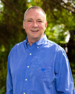 John Lagimodiere has close-cropped grey hair and stands outside wearing a blue button-up collared shirt. He smiles at the camera.