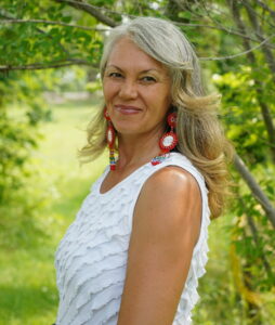 Leah Dorion stands outside in front of a lush green tree. She stands with her left side toward the camera and her head turned to look at the camera as she smiles. Her medium length blonde and grey hair hangs in loose curls and she wears a white sleeveless blouse and red and blue beaded earrings.