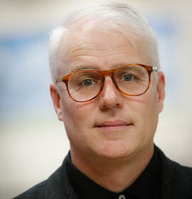 Luke Muller has short, white hair and wears orange-framed glasses. He is wearing a black collared shirt.