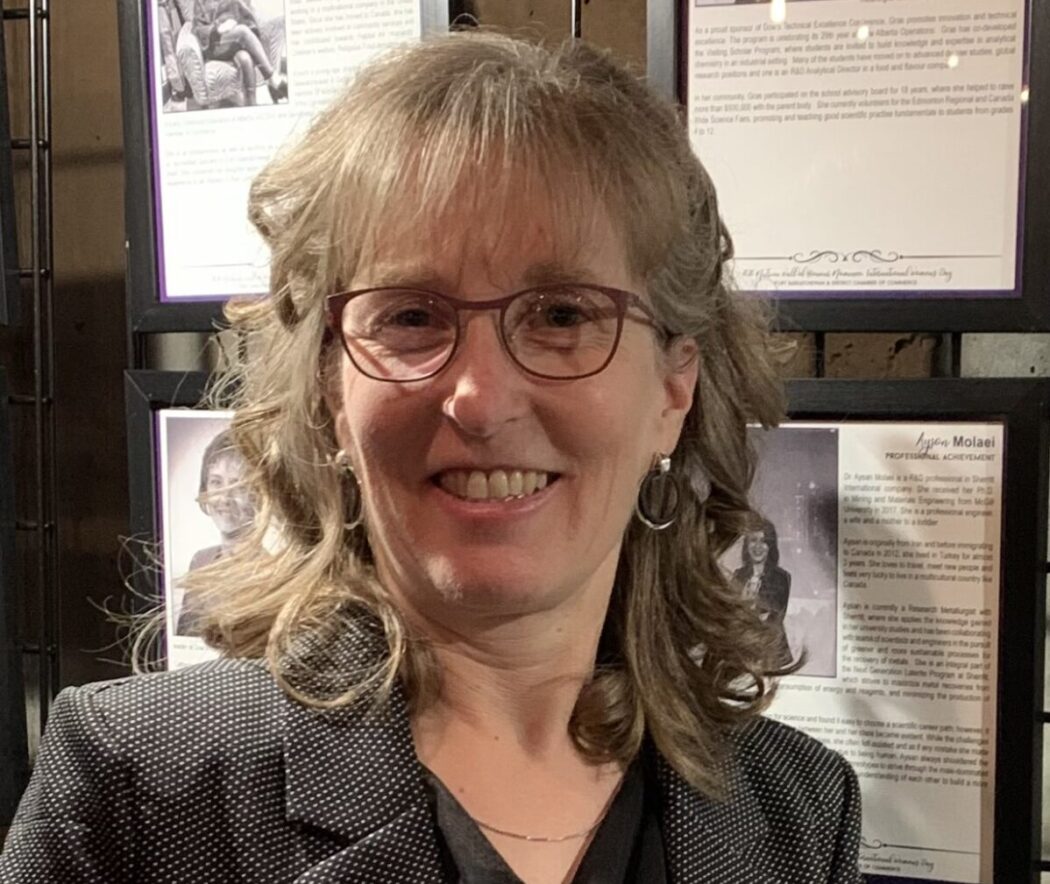 Michele has curly blonde hair that falls just past her shoulders and has glasses. She is wearing a grey blazer with a black shirt underneath.