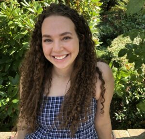 Mikaela Leblanc has long, curly brown hair and is wearing a dark blue and white striped top. She is standing outside in front of some trees and smiling at the camera.