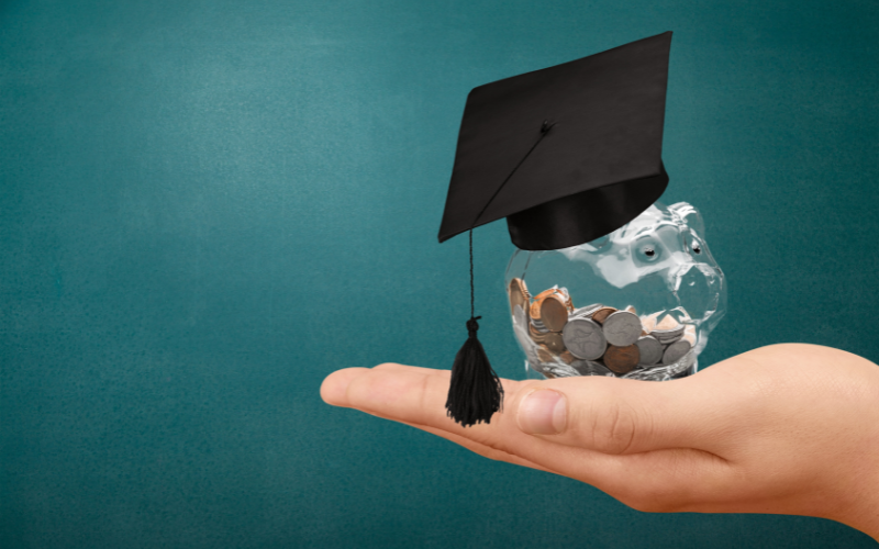 A hand holds a clear plastic piggy bank wearing a graduation hat. The piggy bank has some coins in it.