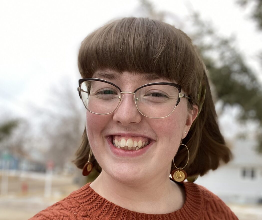 Morgan Kelly wears large glasses and a burnt orange knit sweater. She has brown hair that hang just above her shoulders and bangs that come nearly to the top of her glasses.
