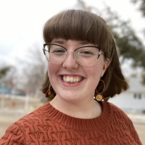 Morgan Kelly wears large glasses and a burnt orange knit sweater. She has brown hair that hang just above her shoulders and bangs that come nearly to the top of her glasses.