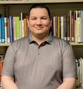 Myles Shingoose has short black hair and is wearing a grey collared T-shirt. He sits in front of a library shelf and smiles at the camera.