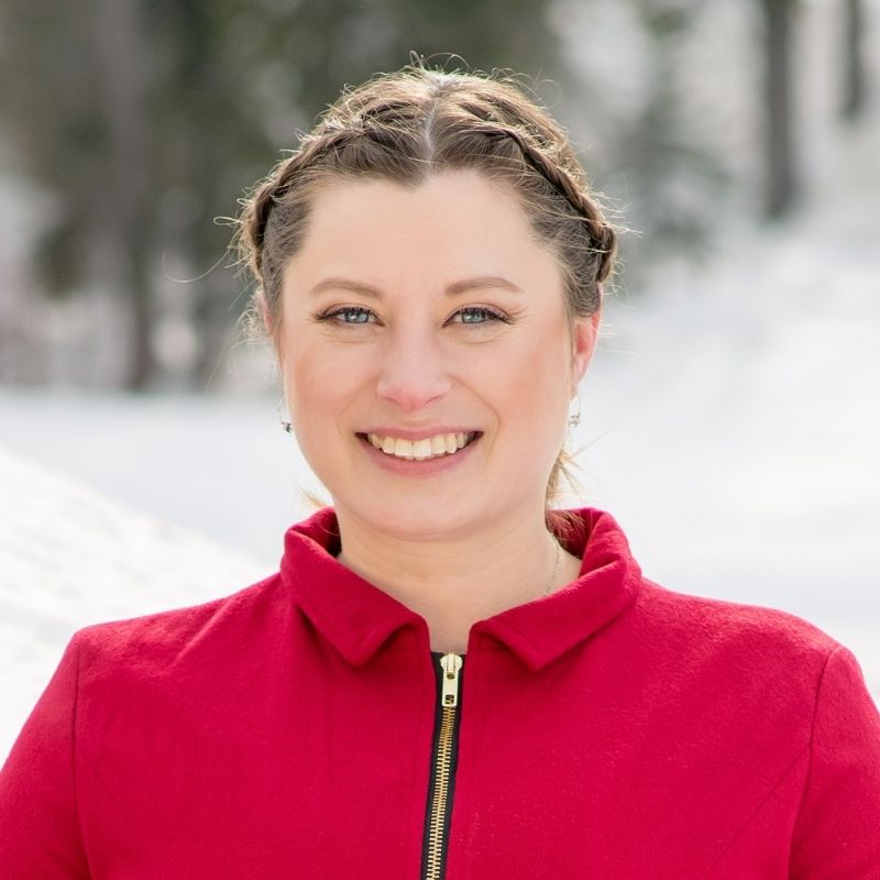 Riane has light brown hair divided into two braids that are pulled back into a bun. She wears a red zip-up sweater with a gold zipper.