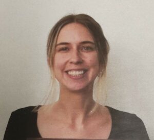 Ruby Lindsay has blonde hair pulled back in a bun and wears a black scoop-neck shirt. She smiles directly at the camera.