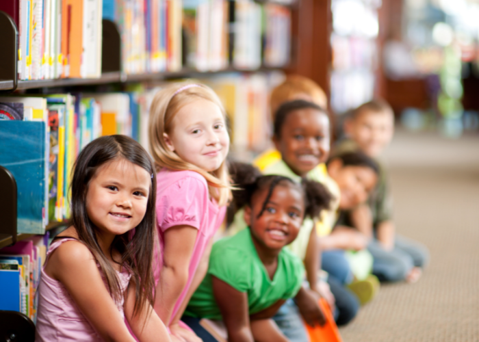 A row of children sit against a bookshelf in a library. The children are looking at the camera and smiling.