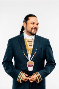 Shawn Cuthand, Writer and Producer at The Feather Entertainment Inc., has long dark hair pulled back into a braid. He wears a dark blue suit and an Indigenous beaded necklace. His head is turned to the left and he smiles at the camera out of the corner of his eye.
