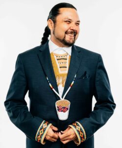 Shawn Cuthand, Writer and Producer at The Feather Entertainment Inc., has long dark hair pulled back into a braid. He wears a dark blue suit and an Indigenous beaded necklace. His head is turned to the left and he smiles at the camera out of the corner of his eye.