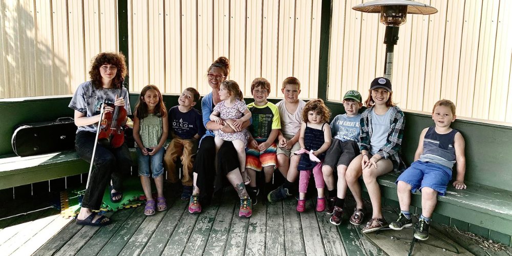 Two adults and nine children sit in an outdoor covered gazebo. The adults are leading a music program for the kids.