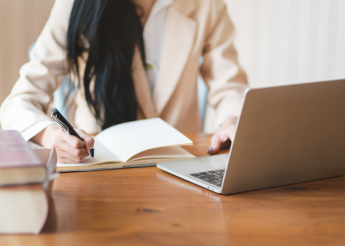 A person sits at a wooden table with a laptop open in front of them while they take notes in a notebook.